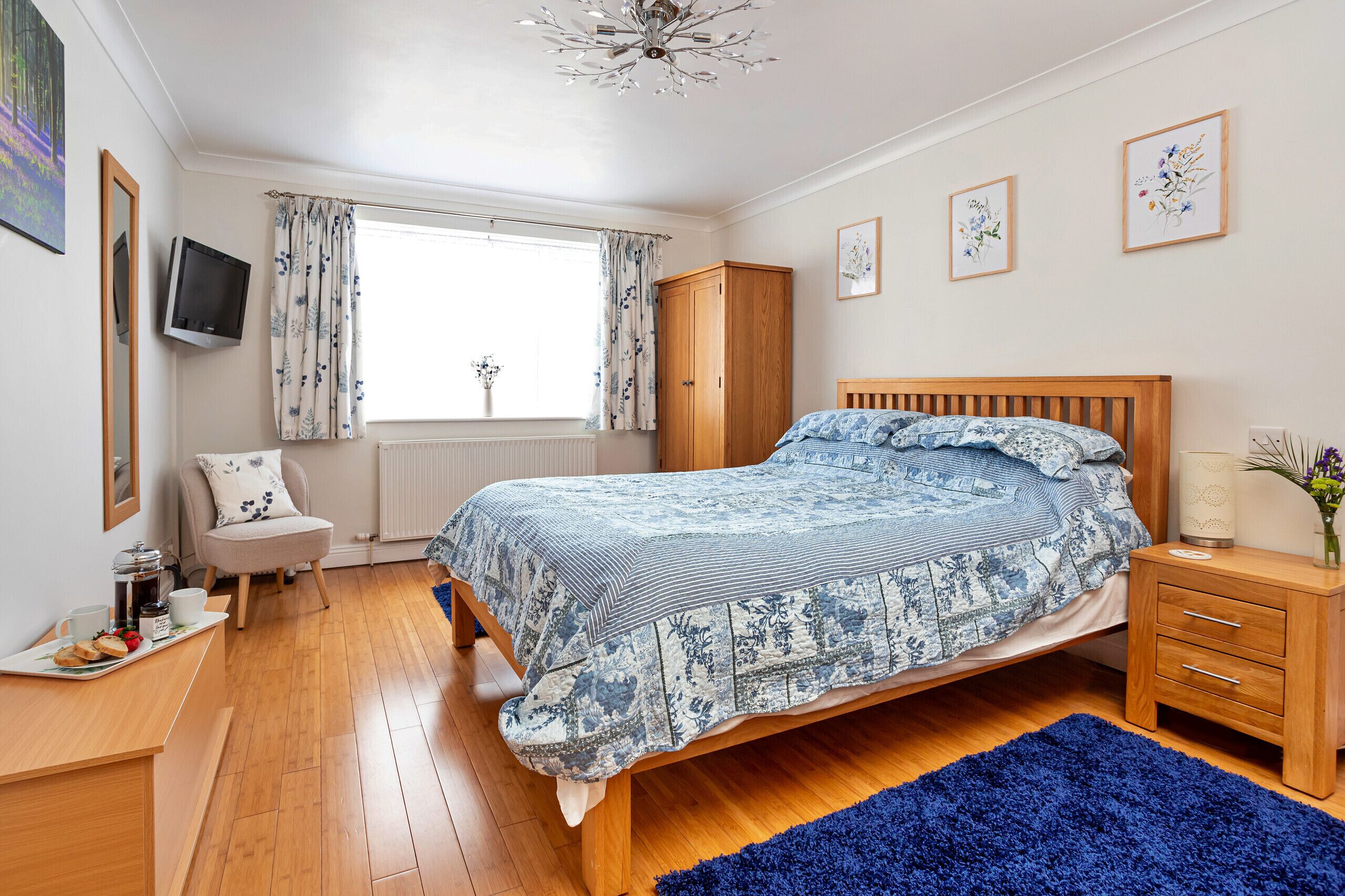 Un dormitorio con cama de madera, mesita de noche, cómoda, televisor montado en la pared y ventana con cortinas.