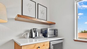 Une petite cuisine équipée avec une machine à café, un four à micro-ondes et un four traditionnel. On y trouve un meuble en bois avec un tiroir, et une étagère au-dessus où sont disposées des reproductions encadrées de motifs botaniques.