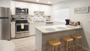 A modern kitchen with white cabinets, stainless steel appliances, a central island with stools, and a 'Beach House' sign on the wall.