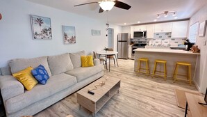 A modern living room with a sofa, coffee table, and a dining area with a table and chairs. A kitchen with a refrigerator, microwave, and bar stools is visible in the background.