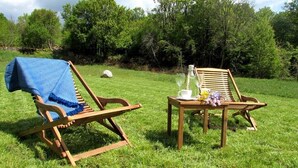 Dos sillones de madera con una mesa entre ellos, ubicados en un campo verde con árboles al fondo.