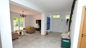 Amplia sala de estar con un sofá, una mesa de centro y un televisor. Hay una puerta que da a otra habitación y una ventana con vistas a la vegetación.