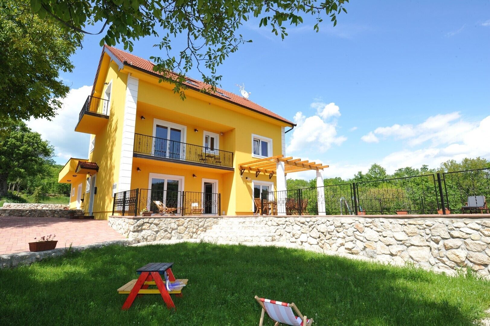 A two-story yellow house with a stone wall in front, a balcony, and a covered patio area.