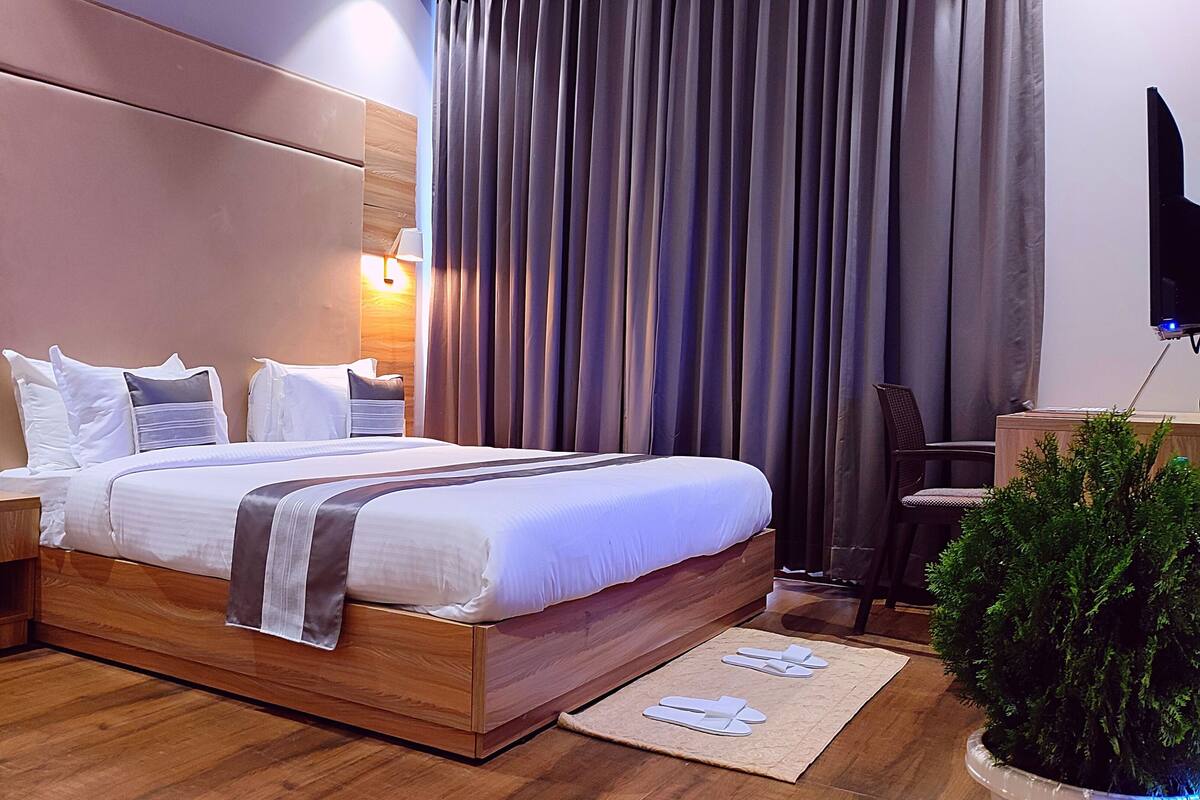 Una moderna camera d'hotel con una testiera in legno, un letto grande con lenzuola bianche, due lampade sui comodini, un televisore a schermo piatto, una scrivania e una pianta in vaso.