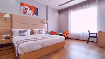 A hotel room with a large bed, a desk, two chairs, a painting on the wall, and a window with curtains.