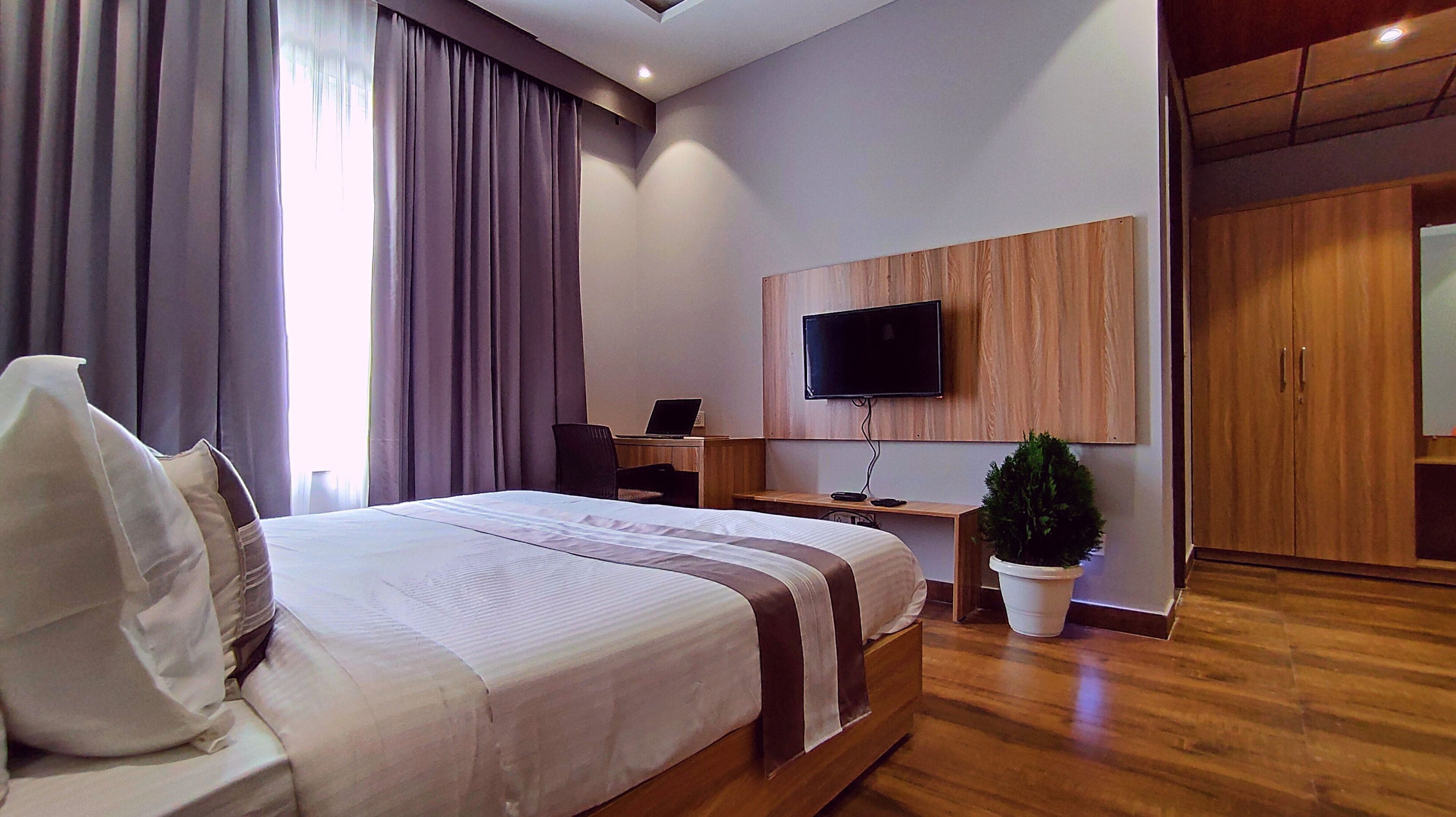 A modern hotel room with a large bed, a desk with a laptop, a flat-screen TV mounted on the wall, and a potted plant.