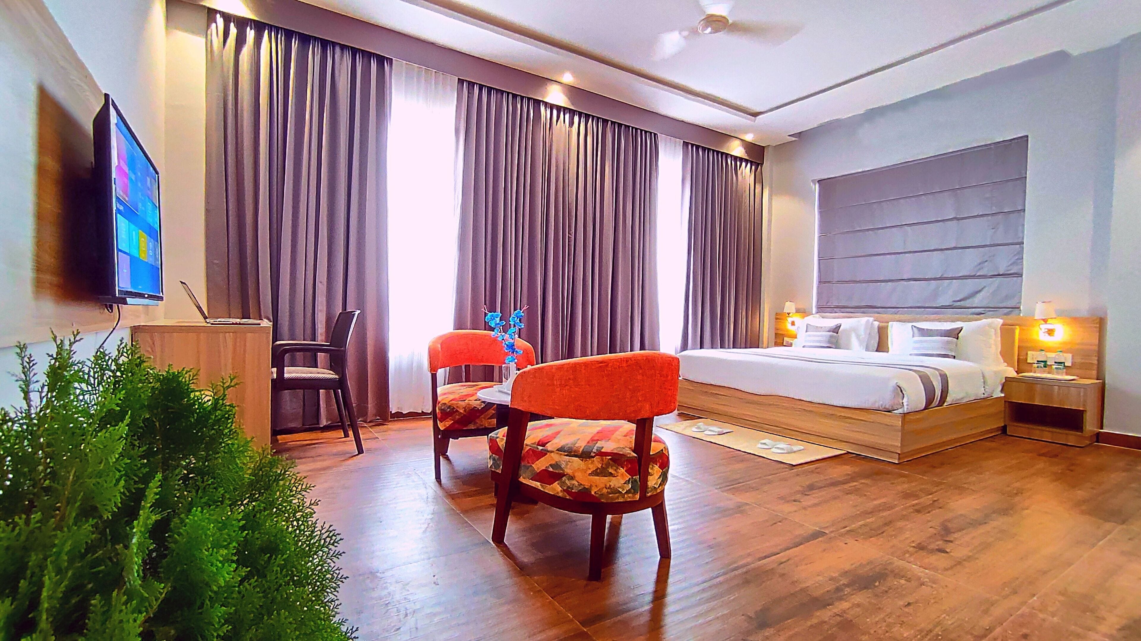 A modern hotel room with a large bed, a desk with a laptop, two chairs, a TV, and a large window with curtains.