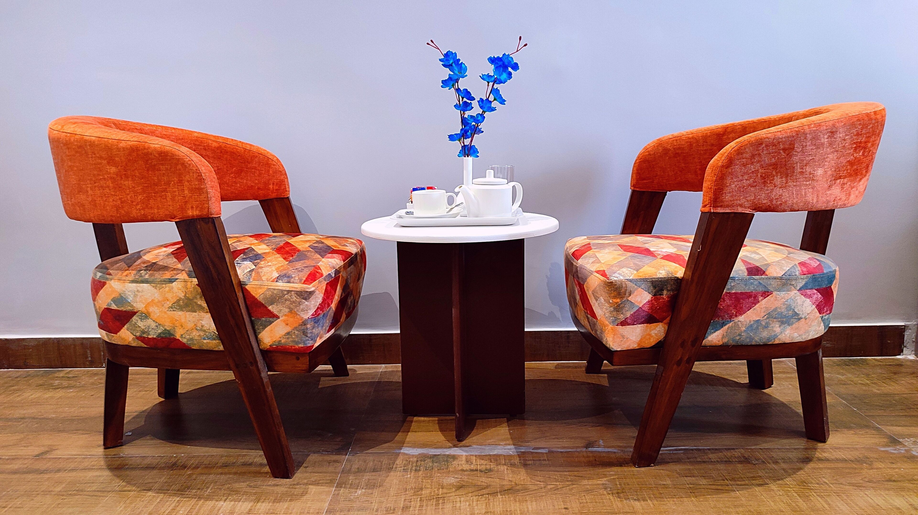 Two chairs with colorful upholstery, a small table with a white vase of blue flowers, and a white teapot and cup on the table.