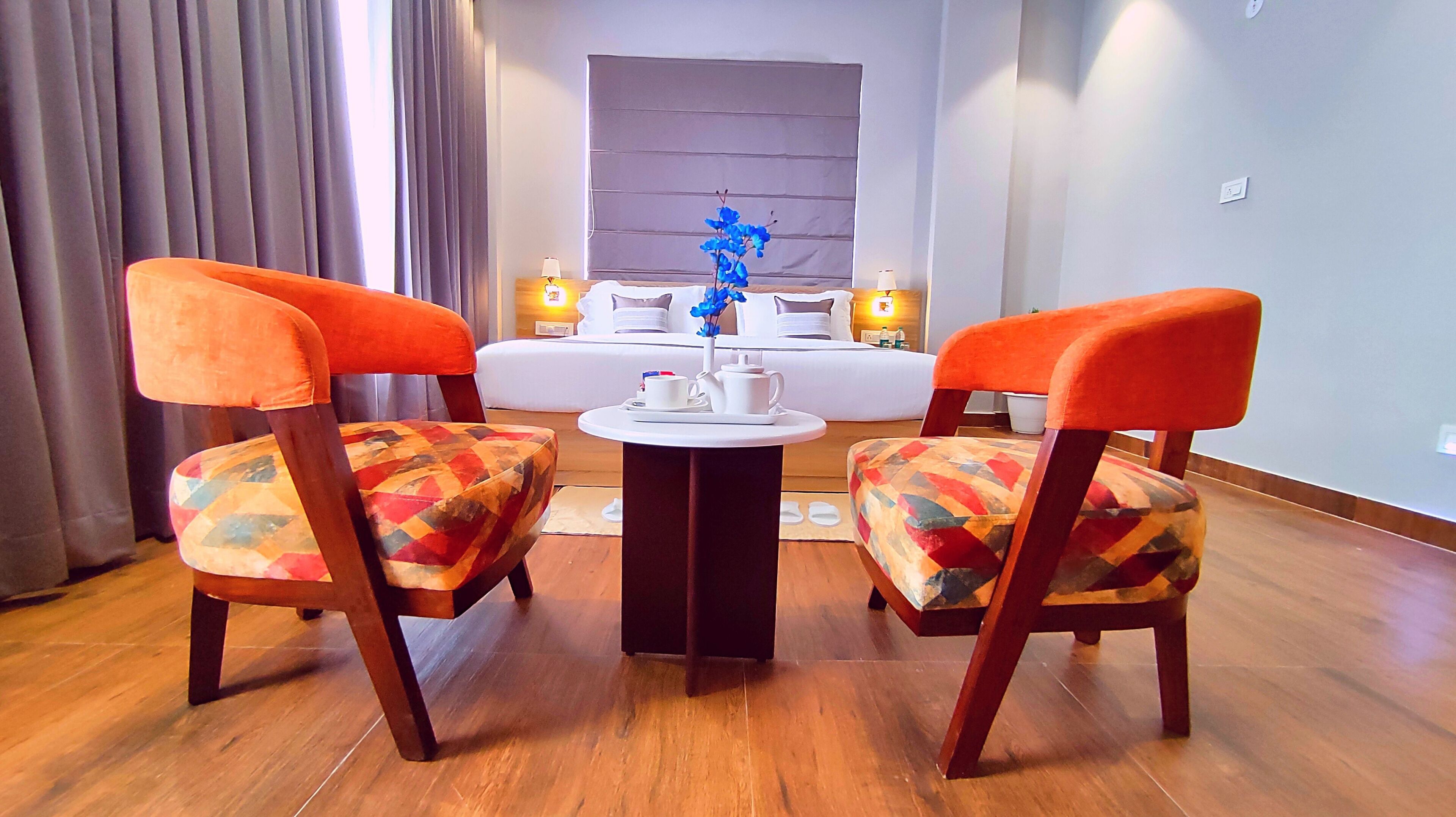A hotel room with two orange chairs, a small round table with a vase of blue flowers, and a bed in the background.