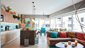 A modern kitchen with a central island, a dining area with a sofa and chairs, and large windows offering a view of the city. - Casa Paraiso - Luxury 2 Bed 3 Bath at Pavilion Lux (Puerto Vallarta)