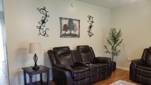 A living room with a dark leather sofa, a small table with a lamp, decorative wall art, and a potted plant. - Spacious 3BR 2BA, 11 Min to Cummins Falls Park (Cookeville)