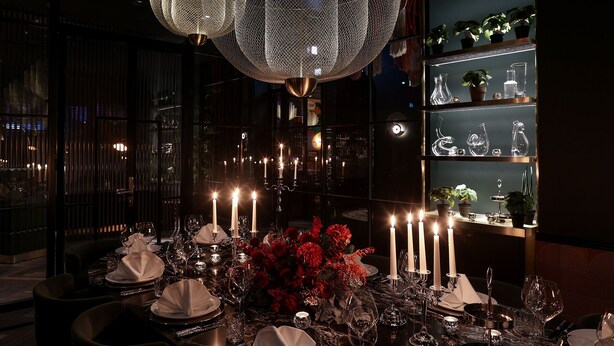 A dimly lit restaurant with a central table set for a meal, surrounded by candles and red flowers. - Hotel ROMY by AMANO (Berlin)