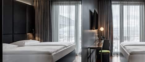 A modern hotel room with two beds, a large window with curtains, a flat-screen TV mounted on the wall, and a small table with a lamp.