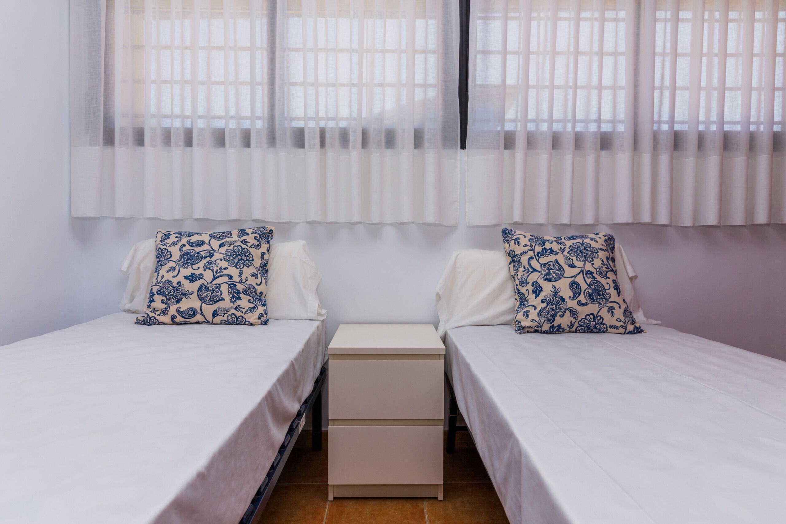 A twin room with two beds, a bedside table, and a window with sheer curtains.