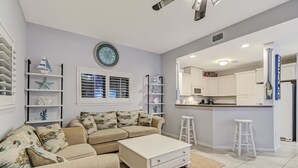 Een woonkamer met een beige bank, een witte salontafel, en een keukenhoek met witte kasten en een magnetron.