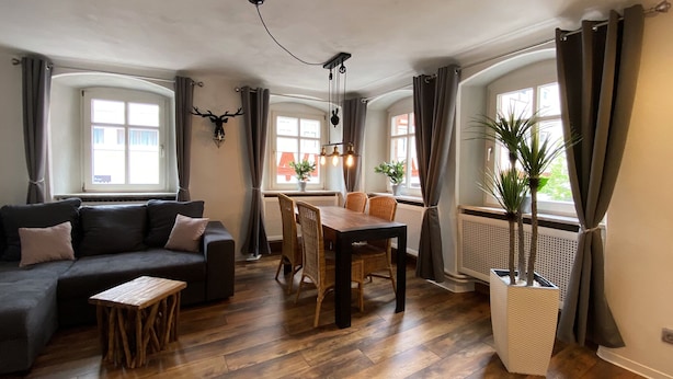 Un salón acogedor con un sofá oscuro, una mesa de centro de madera y una zona de comedor con una mesa de madera y sillas de mimbre.