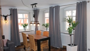 Un comedor con mesa de madera, sillas de mimbre, cortinas grises y plantas en macetas.