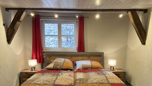 Un dormitorio con una cama de madera, dos mesitas de noche con lámparas, una ventana con cortinas rojas y un techo inclinado.