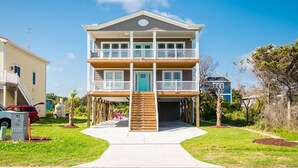 A two-story building with a balcony on the upper floor, a wooden staircase leading up to the entrance, and a clear sky in the background. - Sweet Sunshine: Lovely Rental with Pool and Elevator Built in 2021 (Oak Island)