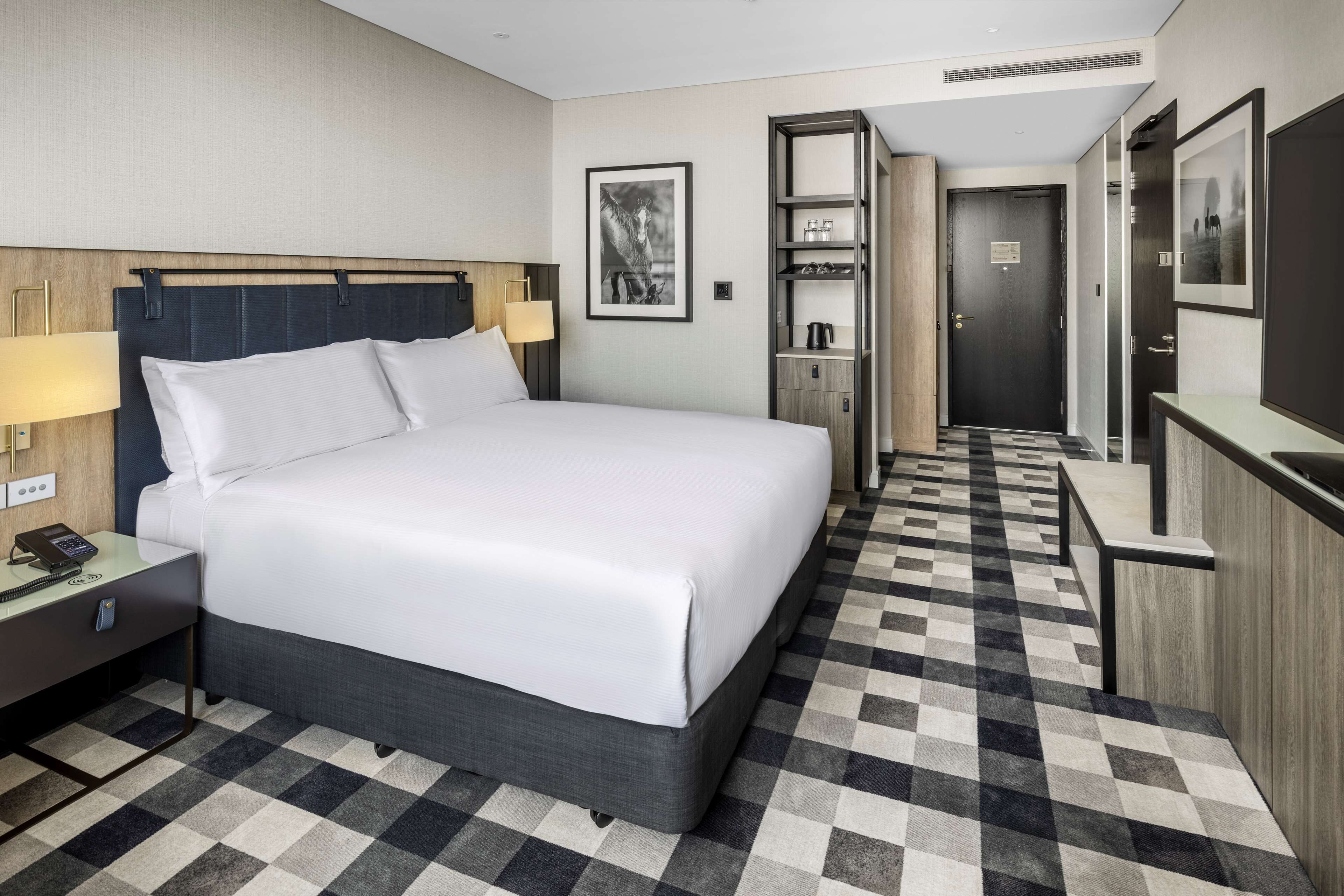 A hotel room with a large bed, a desk with a lamp, a wardrobe, and a bathroom visible through an open door.