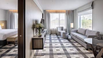 Habitación de hotel con cama, sofá, mesa de comedor, mesita auxiliar y ventana grande con cortinas.