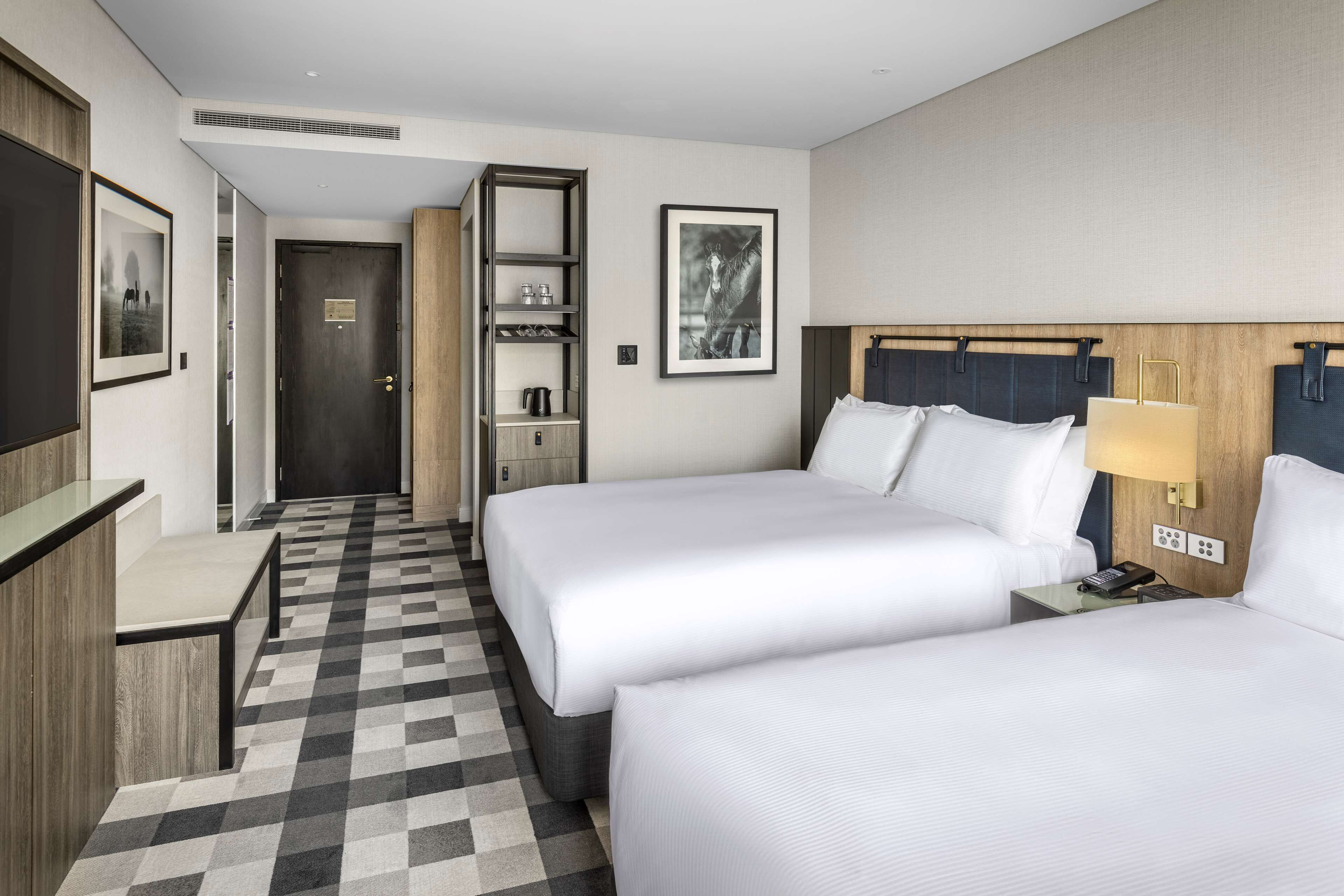 Habitación de hotel con dos camas, un escritorio, una silla, un armario, una estantería con objetos y un cuadro en la pared.