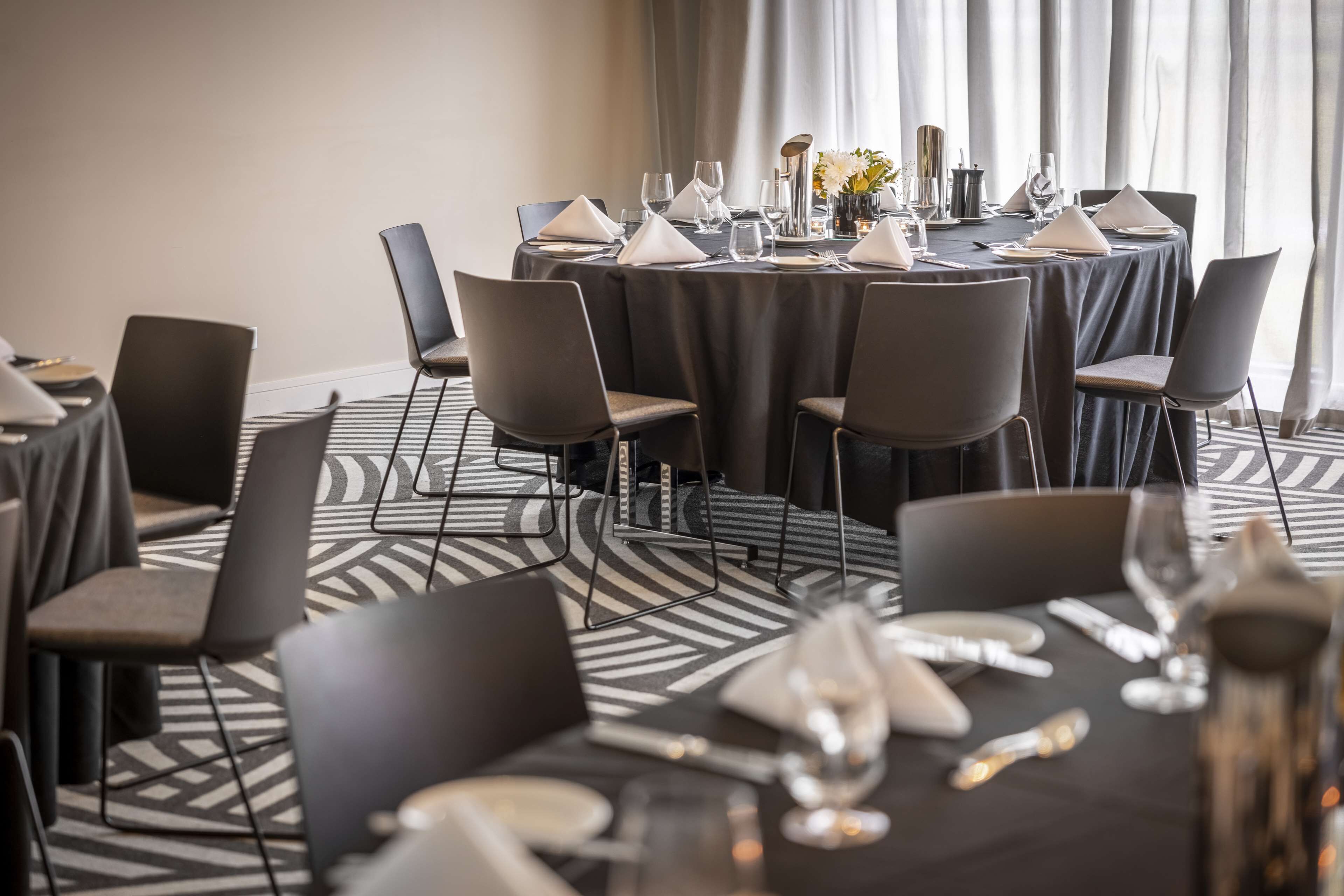 A dining area with round tables set for a meal, each with a white tablecloth, black chairs, and neatly arranged cutlery and glassware.