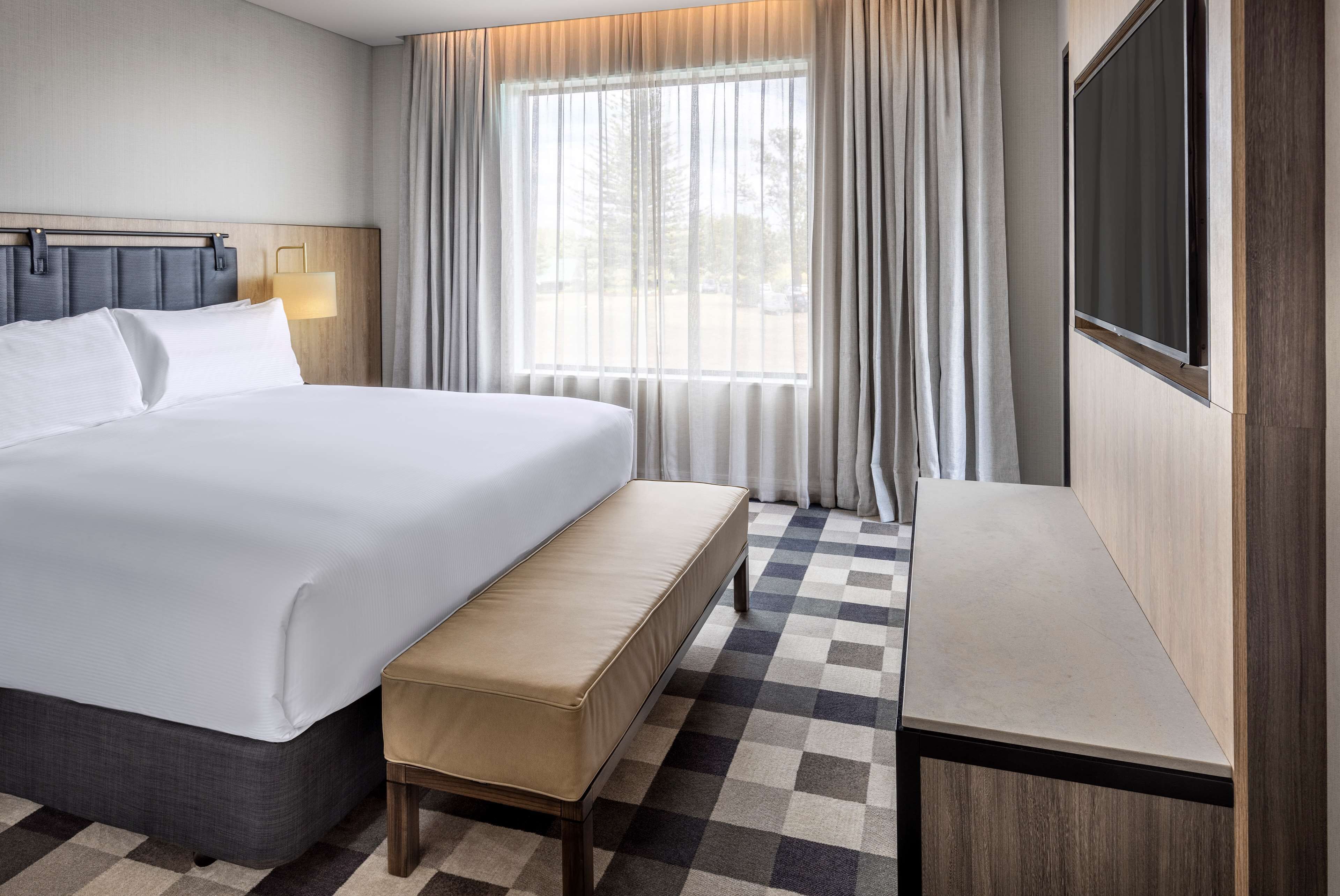 A hotel room with a large bed, a bench, a desk, and a large window with curtains.