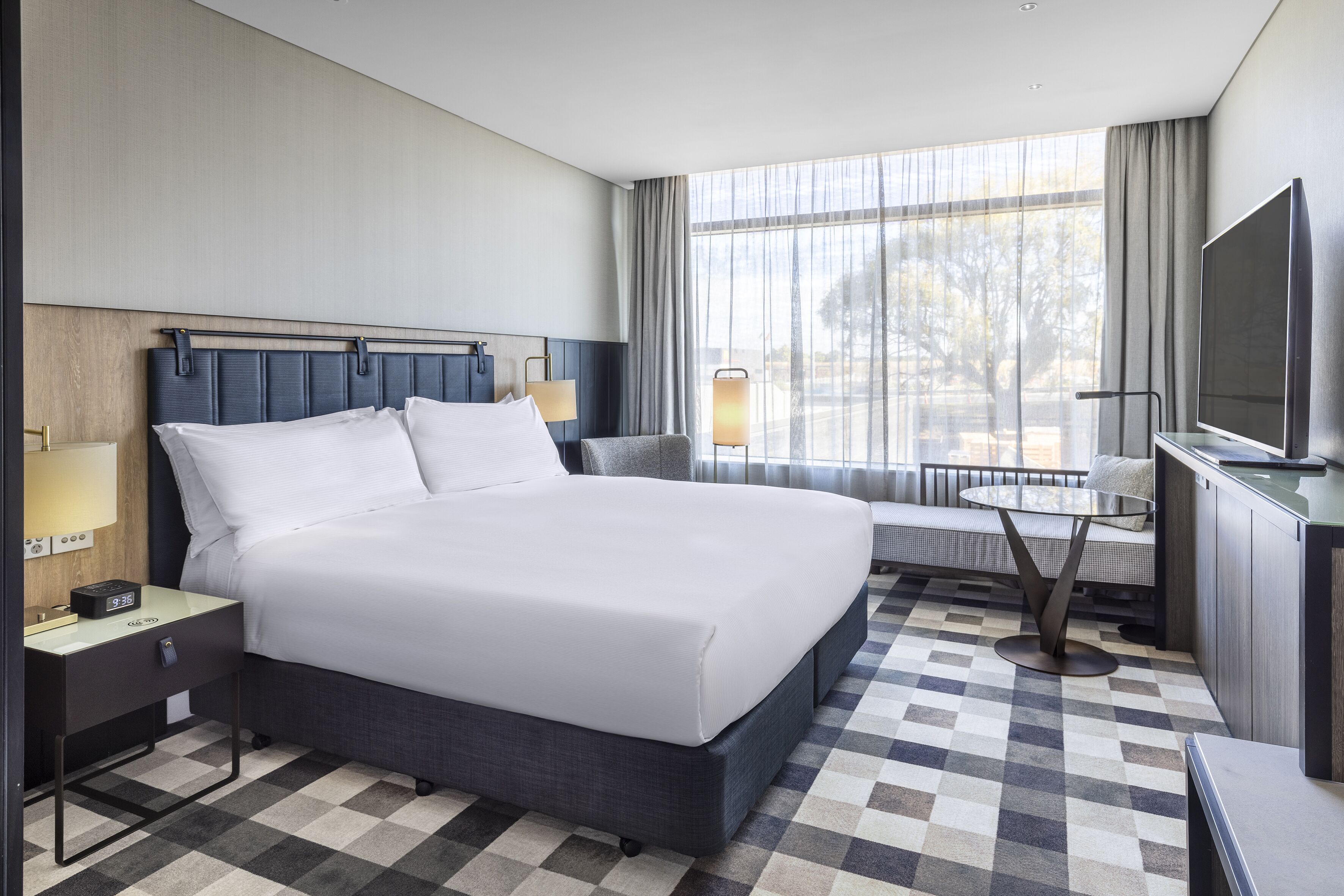 Una habitación de hotel moderna con una cama grande, mesitas de noche, un televisor y vistas al exterior.