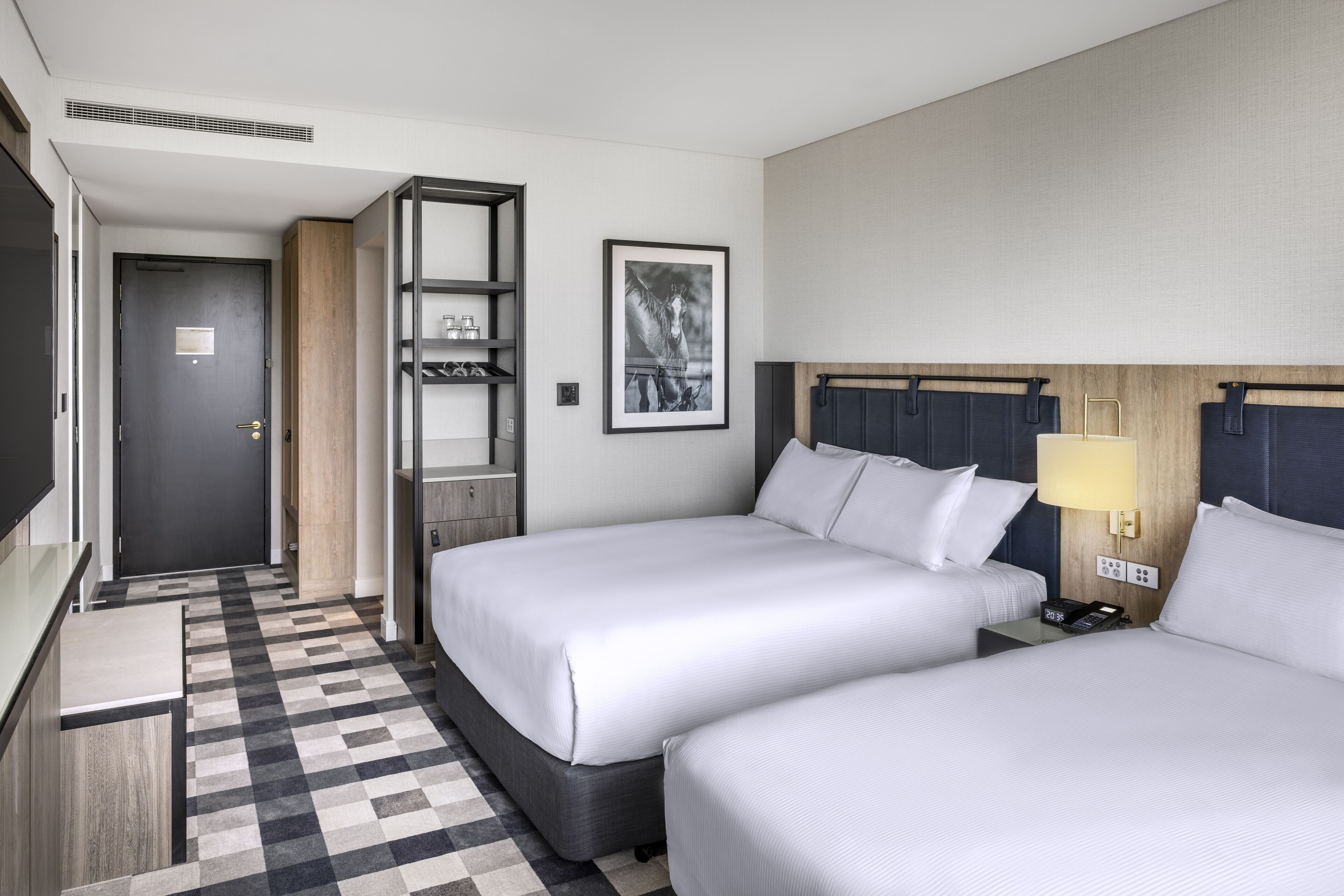 A hotel room with two beds, a check-in counter, a coat rack, a framed picture, and a lamp.
