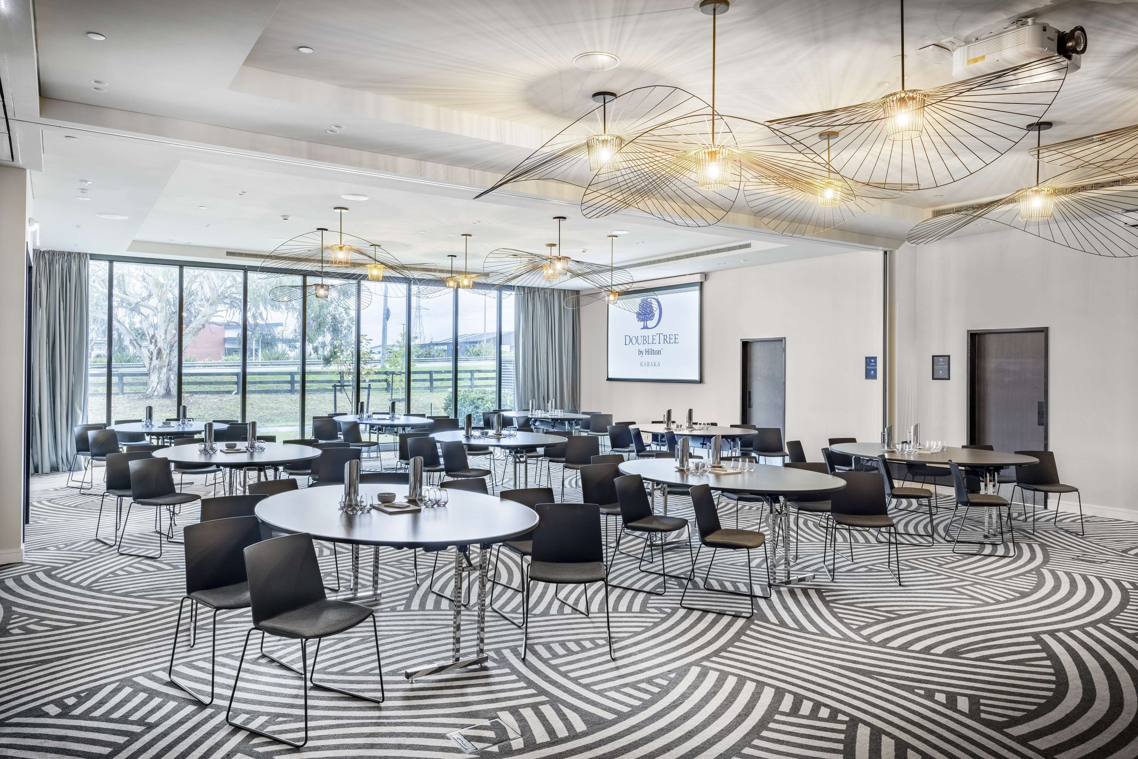A conference room with round tables and chairs, large windows, and a projector screen displaying "DOUBLETREE by Hilton".