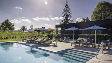 Une piscine entourée de verdure et d’un bâtiment moderne, avec des chaises longues et des parasols à proximité.