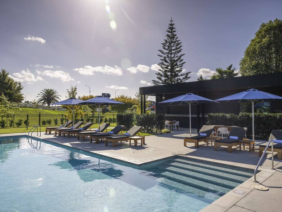 Une piscine entourée de verdure et d’un bâtiment moderne, avec des chaises longues et des parasols à proximité.