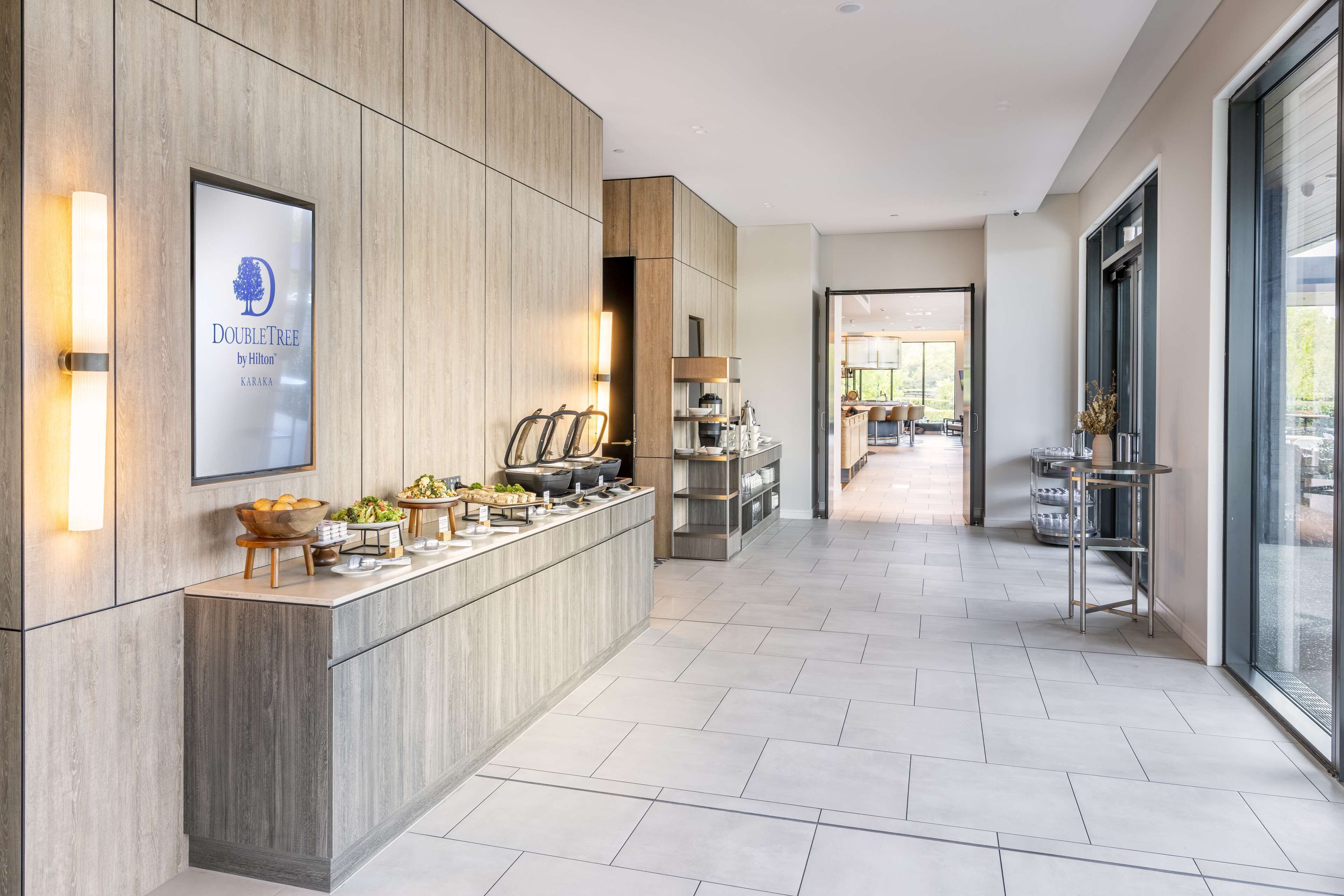 Een moderne hotellobby met een centrale balie, een lijst met de tekst 'DoubleTree by Hilton KARA', en uitzicht op de eetruimte.
