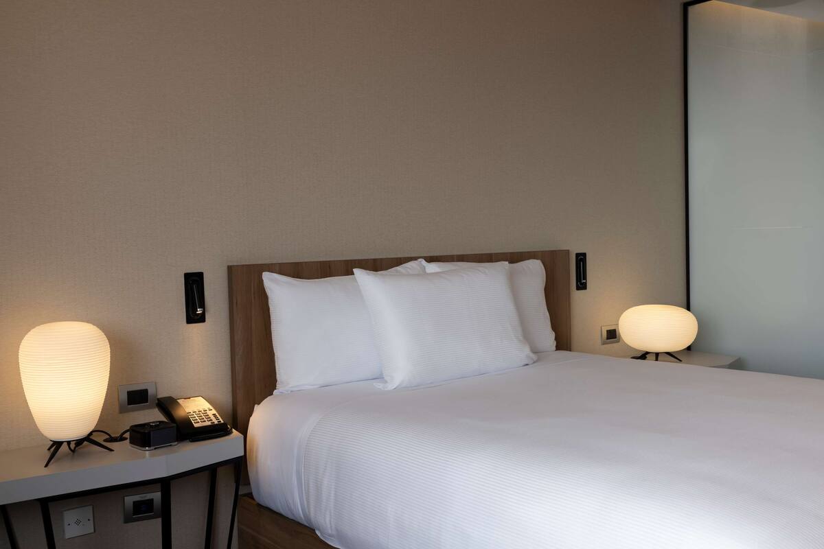 A neatly made bed with white linens, a bedside lamp, a phone, and a wall-mounted light fixture in a hotel room.
