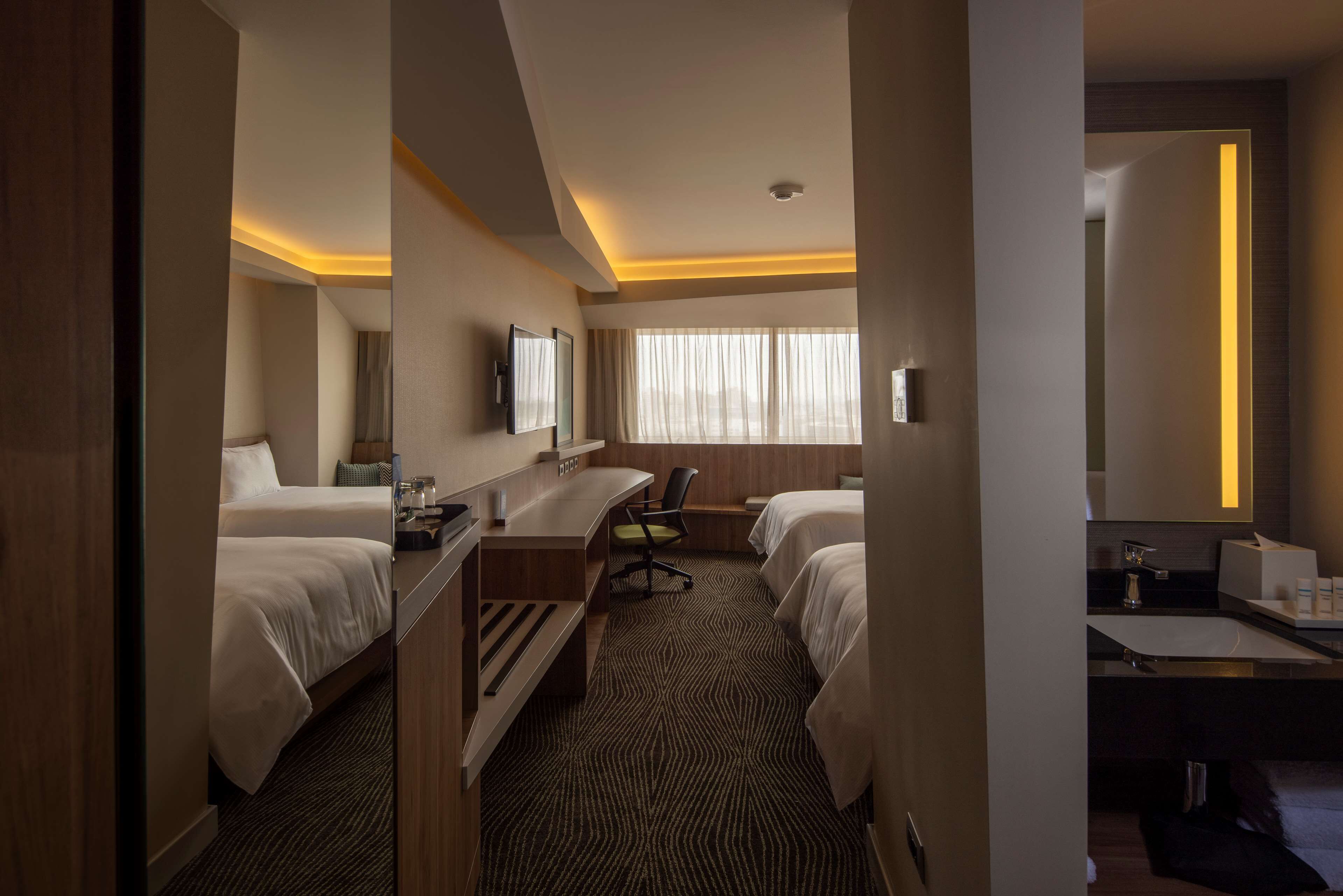 A hotel room with two beds, a desk, a chair, a large mirror, and a window with curtains.