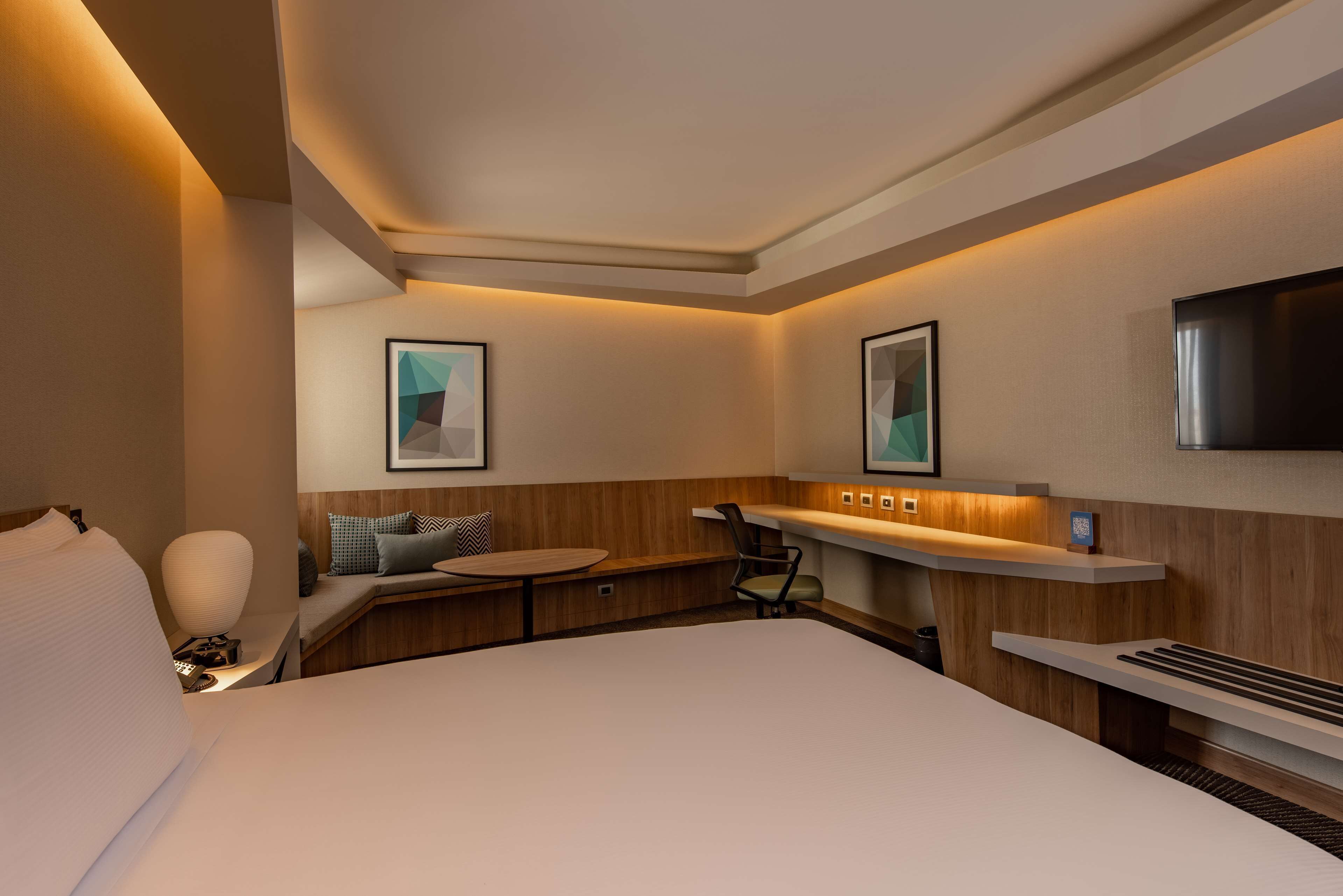 Una habitación de hotel moderna con una cama grande, un escritorio, una silla, una mesita, un televisor y obras de arte en las paredes.