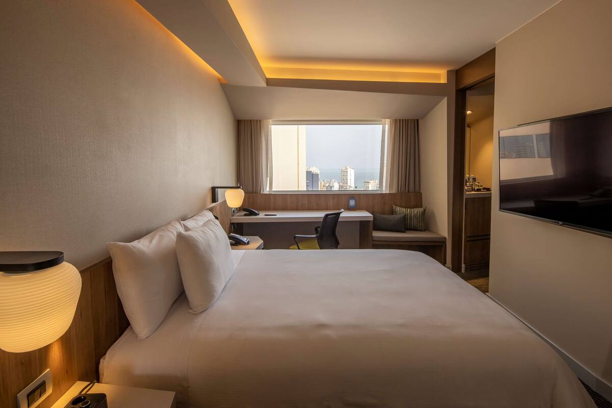 A hotel room with a large bed, a desk, a chair, a TV, and a window with a city view.