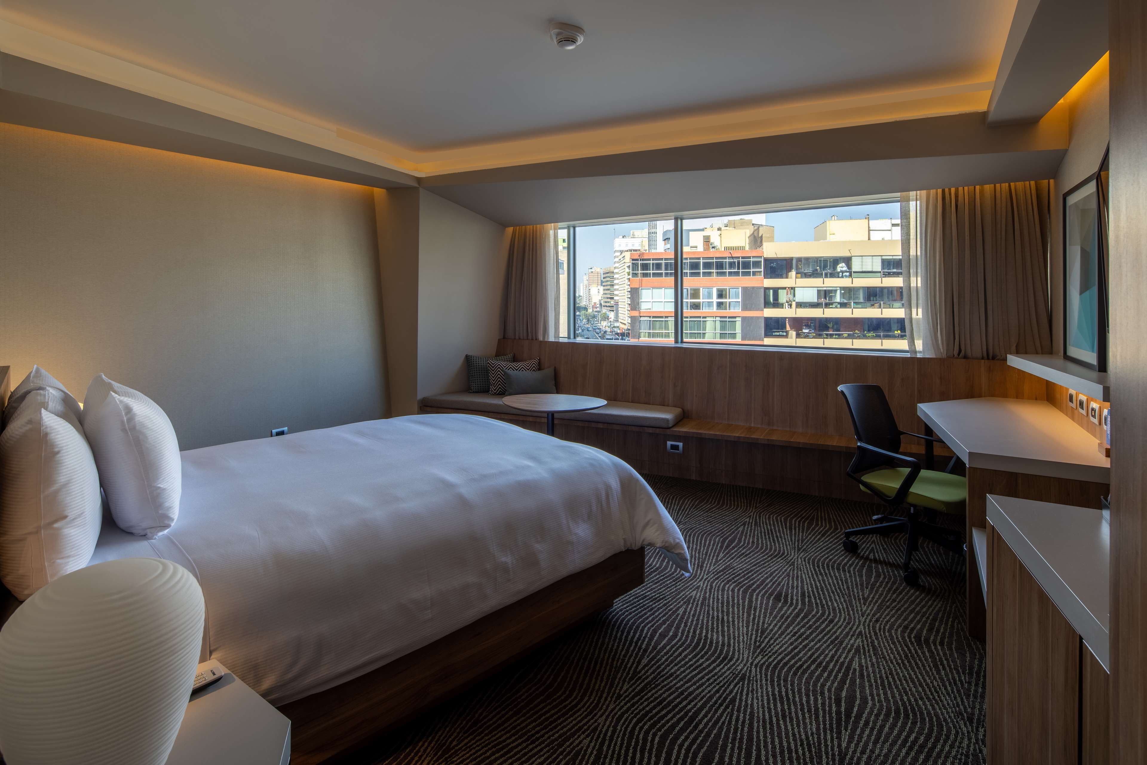 A hotel room with a large bed, a desk, a chair, a small round table, and a view of the city through a large window.