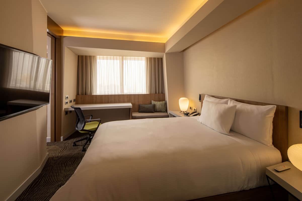 A hotel room with a large bed, a desk with a chair, a TV, a window with curtains, and a bedside lamp.
