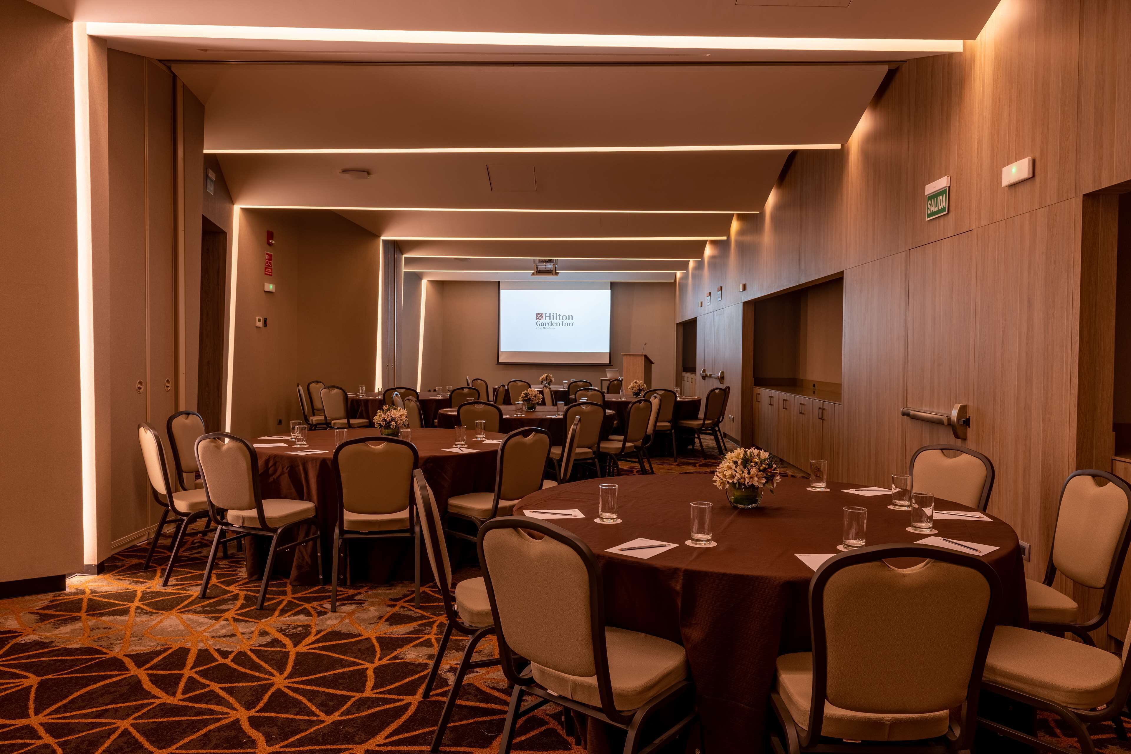 A conference room with multiple tables and chairs, a projector screen displaying a presentation, and a carpeted floor.