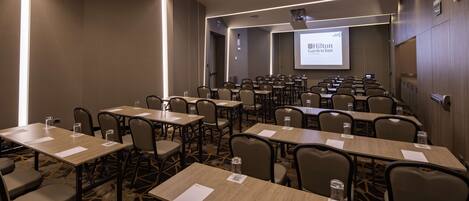 A conference room with multiple tables and chairs, a projector screen displaying 'Hilton Garden Inn', and a carpeted floor.