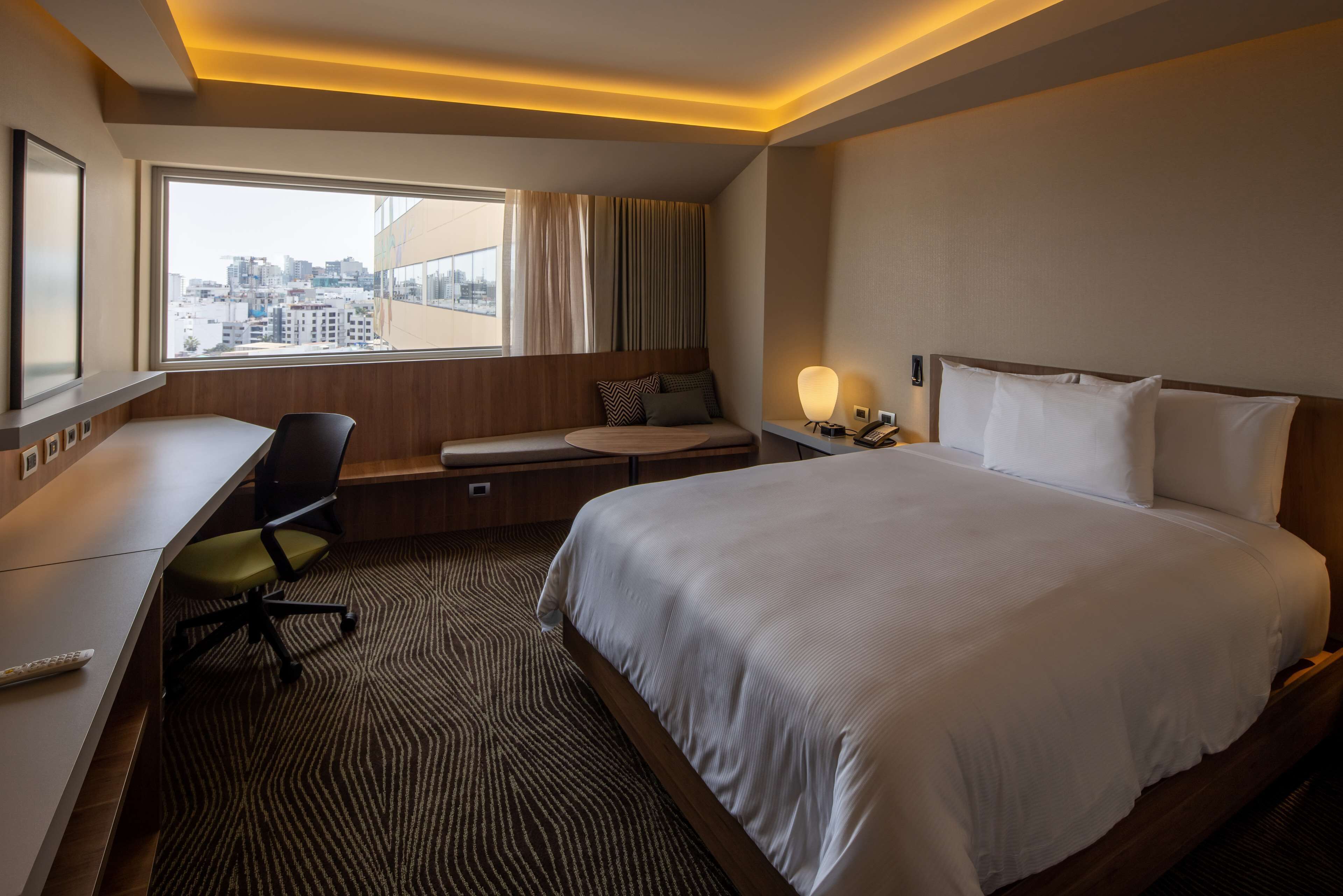 Una habitación de hotel con cama, escritorio, silla y vistas de la ciudad a través de un amplio ventanal.