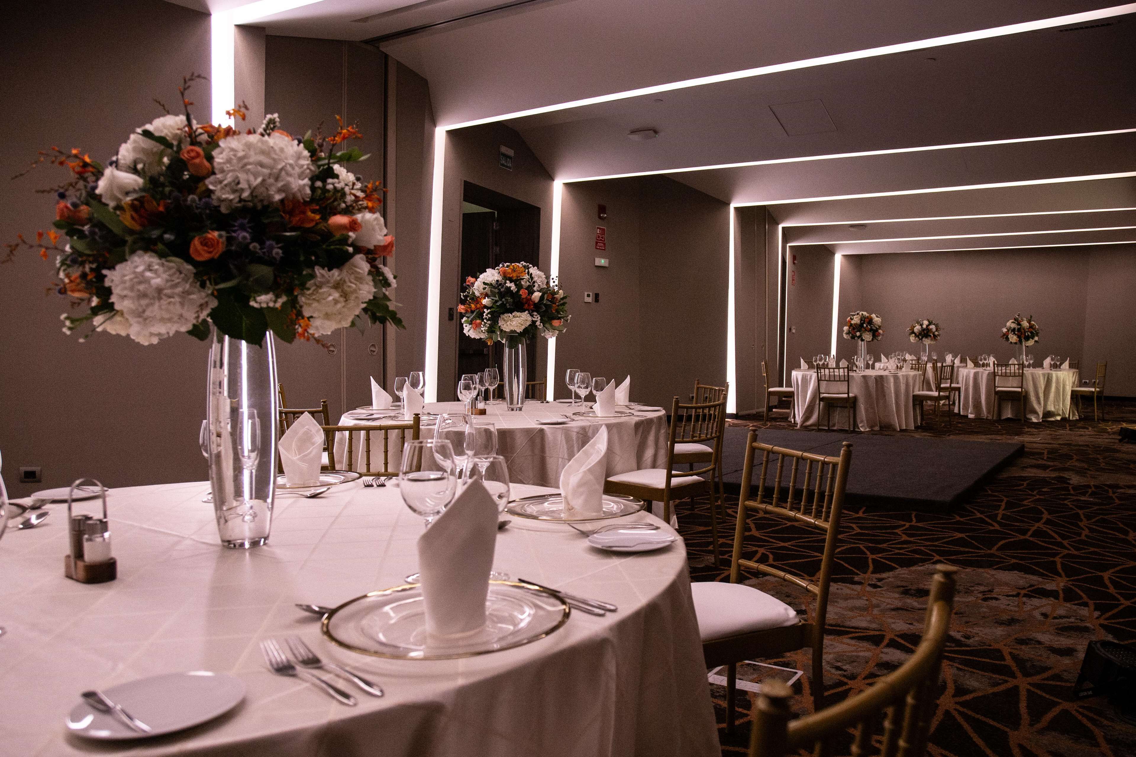 A banquet hall with round tables set for dining, each with white tablecloths, glassware, and floral centerpieces.