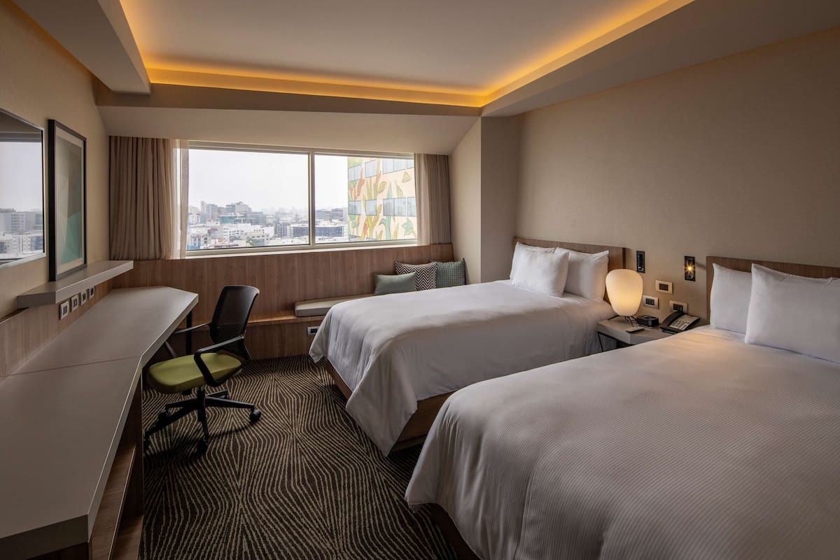 A hotel room with two beds, a desk, a chair, a window with a city view, and a patterned carpet.
