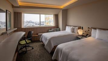 Quarto de hotel com duas camas, uma escrivaninha, uma cadeira, janela com vista para a cidade e tapete estampado.