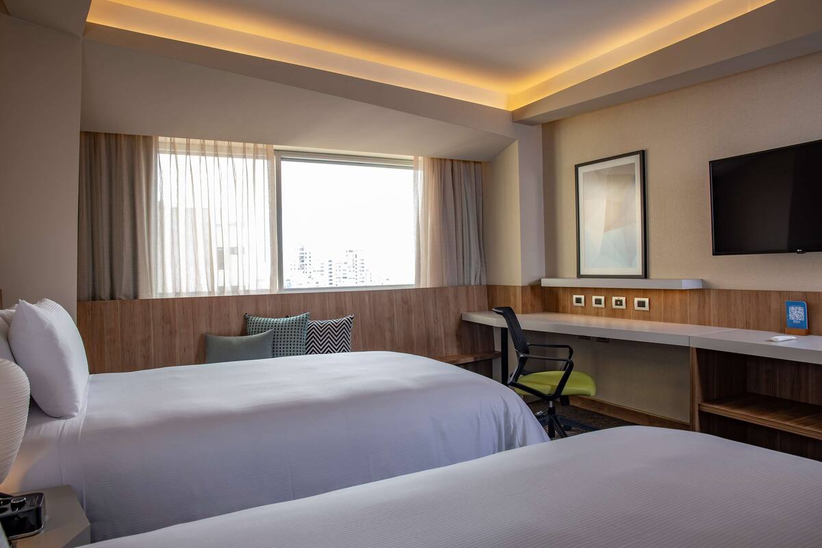 A hotel room with two beds, a desk, a chair, a TV, and a window with curtains.