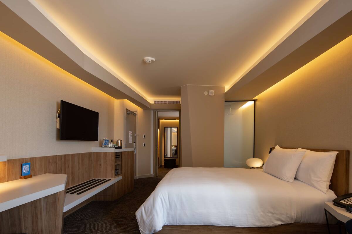 A hotel room with a bed, a TV mounted on the wall, a bedside table with a lamp, and a reception desk with a counter.
