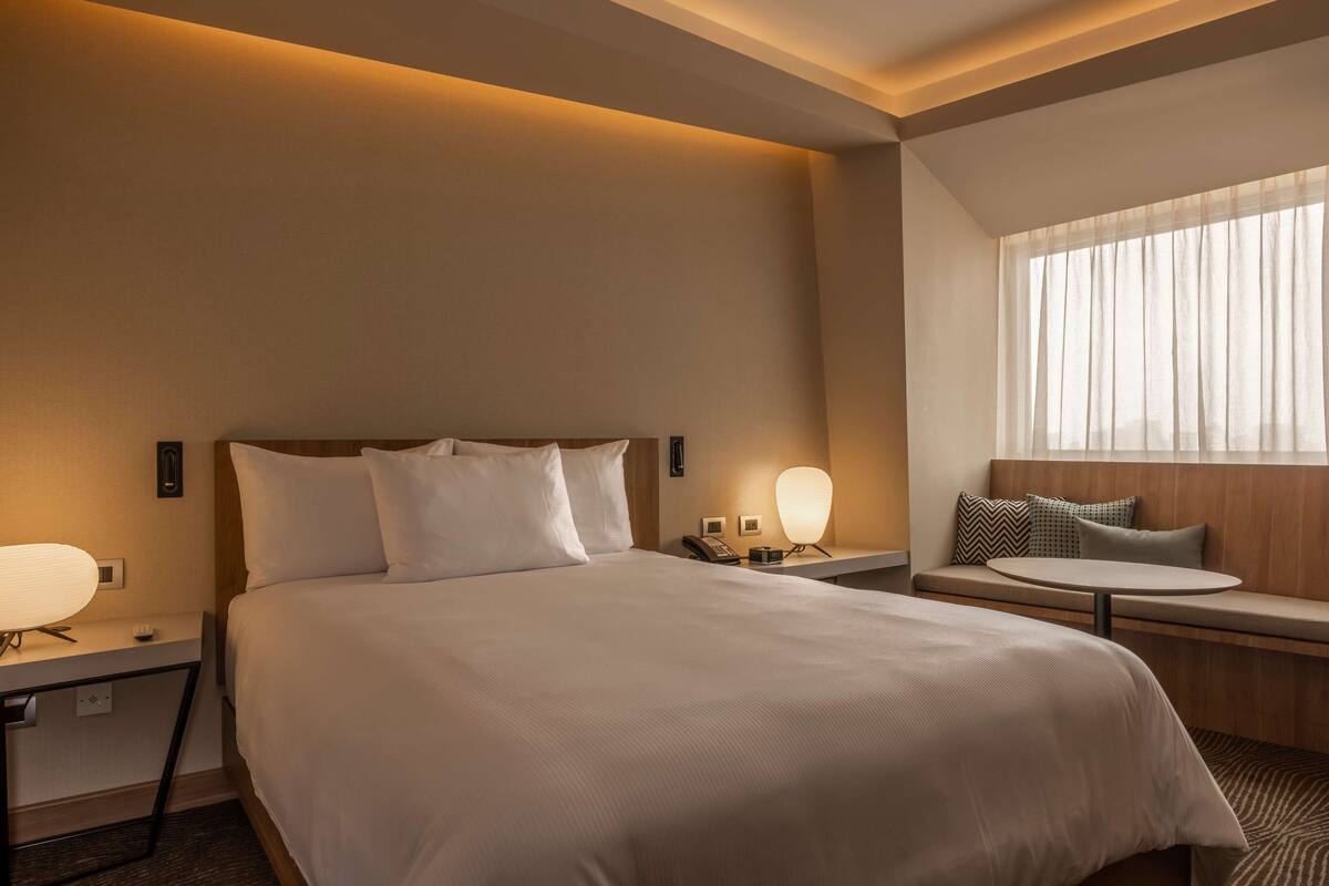 A hotel room with a large bed, bedside tables with lamps, a small round table, and a bench with cushions.