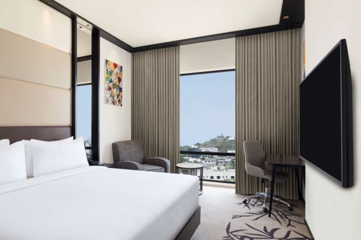 A hotel room with a large bed, a desk, a chair, a TV, and a view of the city through large windows.
